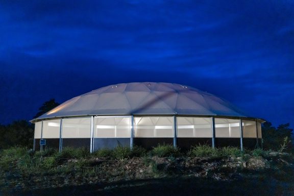Longierhalle bei Nacht Runde, moderne Struktur mit beleuchteten Fenstern unter einem dunklen, bewölkten Himmel.