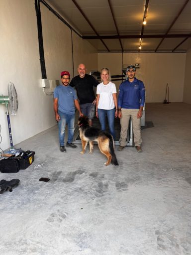 Vier Personen stehen in einem leeren Raum, ein Schäferhund ist ebenfalls anwesend.