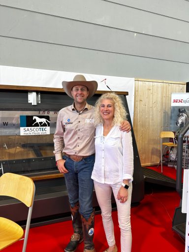 Grischa Ludwig Mann im Cowboy-Hut und Frau in weißem Shirt stehen lächelnd vor einem Ausstellungsstand.